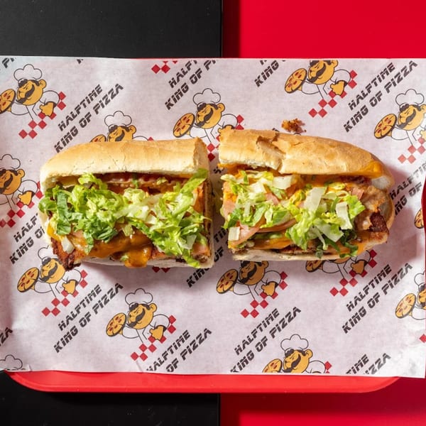 A Halftime Pizza sub sandwich, cut in half, with lettuce, sauce, and meat on branded paper.
