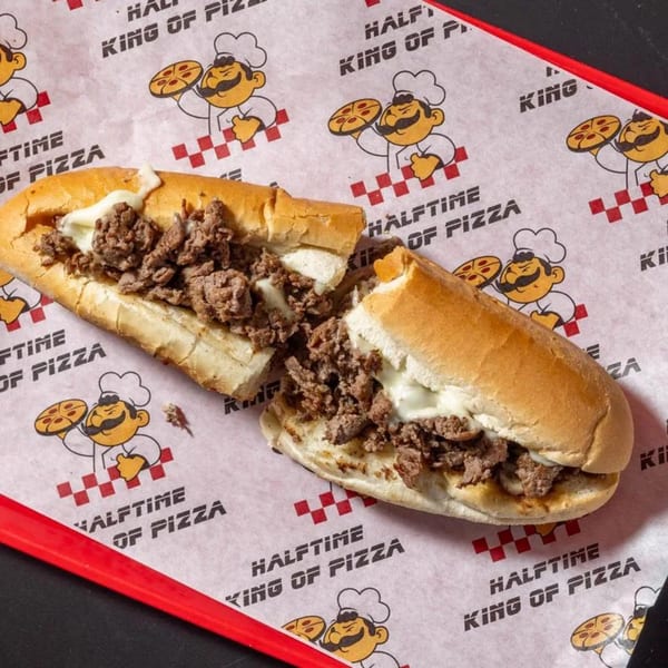 Halftime Pizza's cheesesteak sandwich, cut in half, on branded wax paper with a red tray.